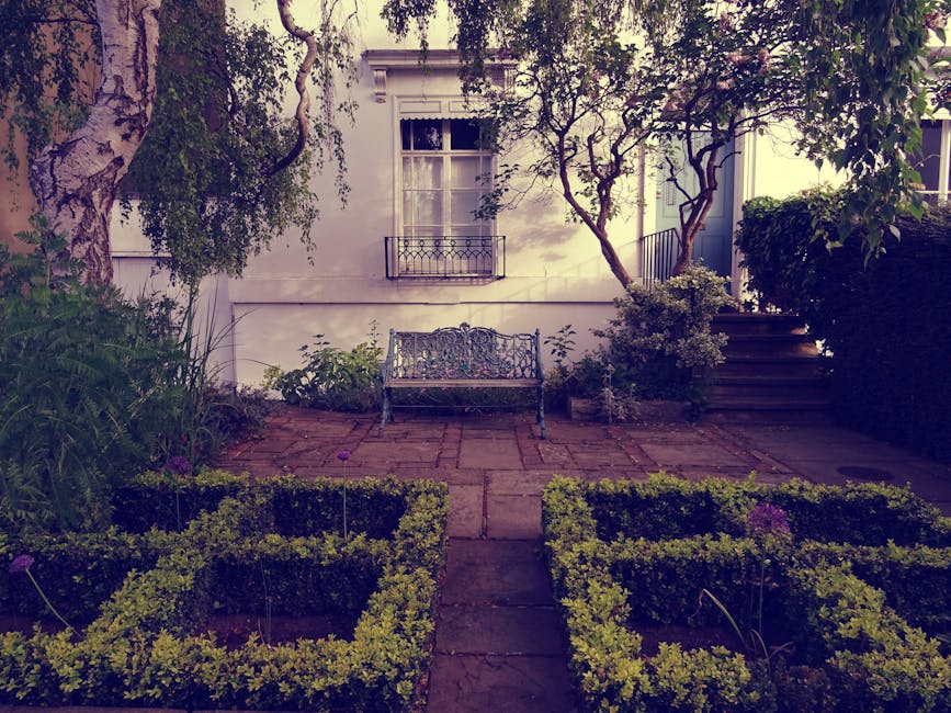 A small, well-maintained garden with a paved stone patio area featuring a decorative cast iron bench centered against a white house wall. To the left, lush green plants and bushes grow beside a large 