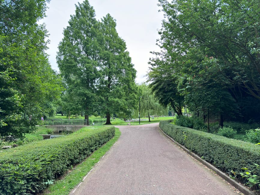 A winding pathway through a lush, well-maintained park featuring neatly trimmed hedges along both sides, tall deciduous trees with full green foliage creating a canopy over the path, and grassy areas 